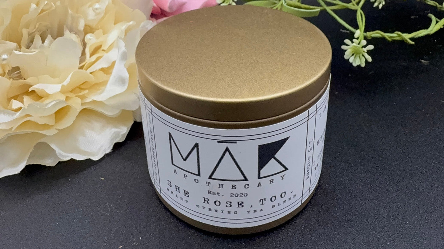 gold can with white lable and black writing that says She Rose , Too. Flowers in back on black background.